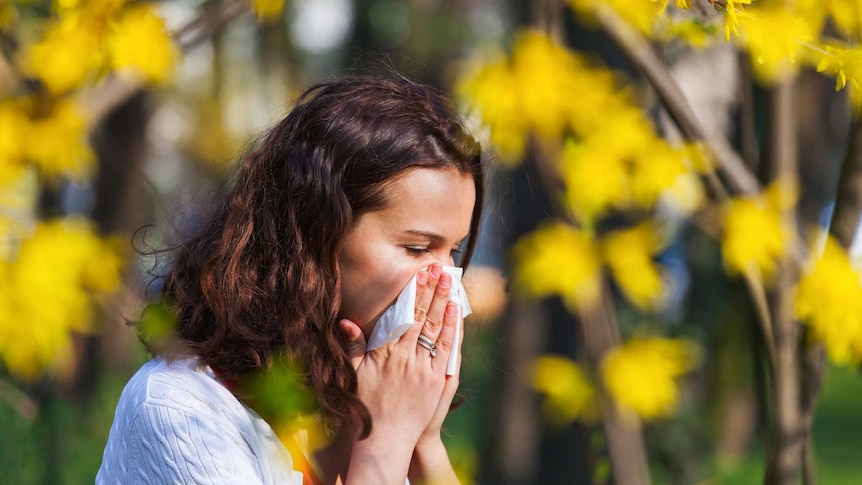 Woman blowing her nose outside, standing in front of yellow flowers