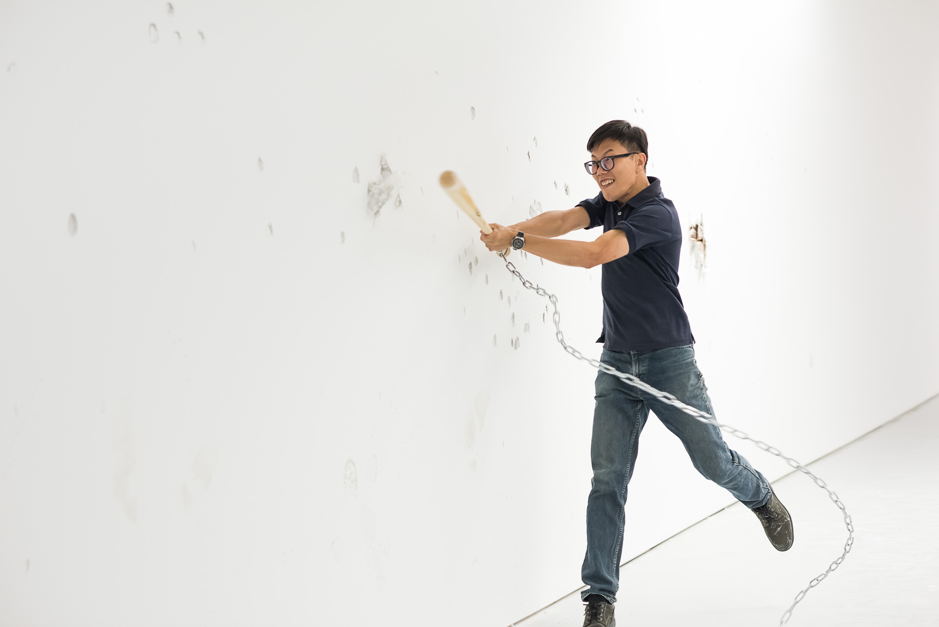 An Asian man uses a baseball bat attached to a chain to hit a white gallery wall. Indents can be seen in the wall.