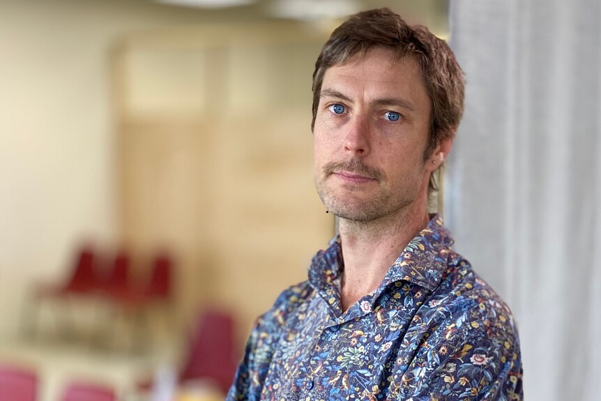 A man in a colourful patterned shirt stands against a wall indoors.