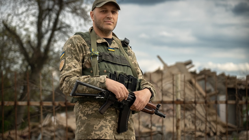 A man wearing a caop and army fatigues holds a gun and backpack.