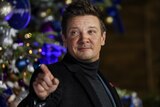 Jeremy Renner smiles and points a finger at something off camera. 