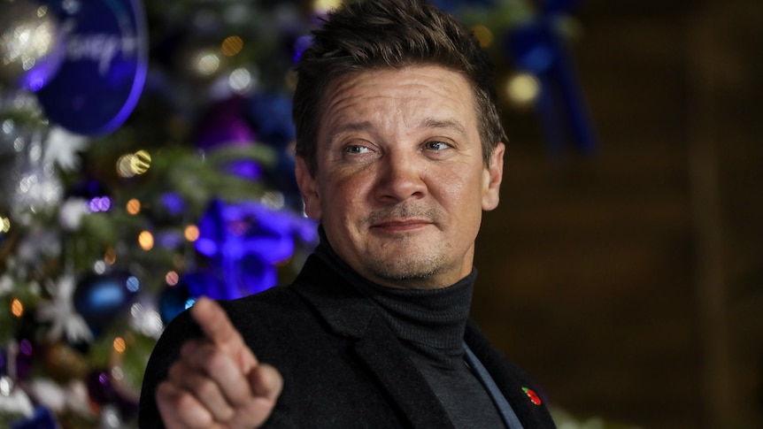 Jeremy Renner smiles and points a finger at something off camera. 