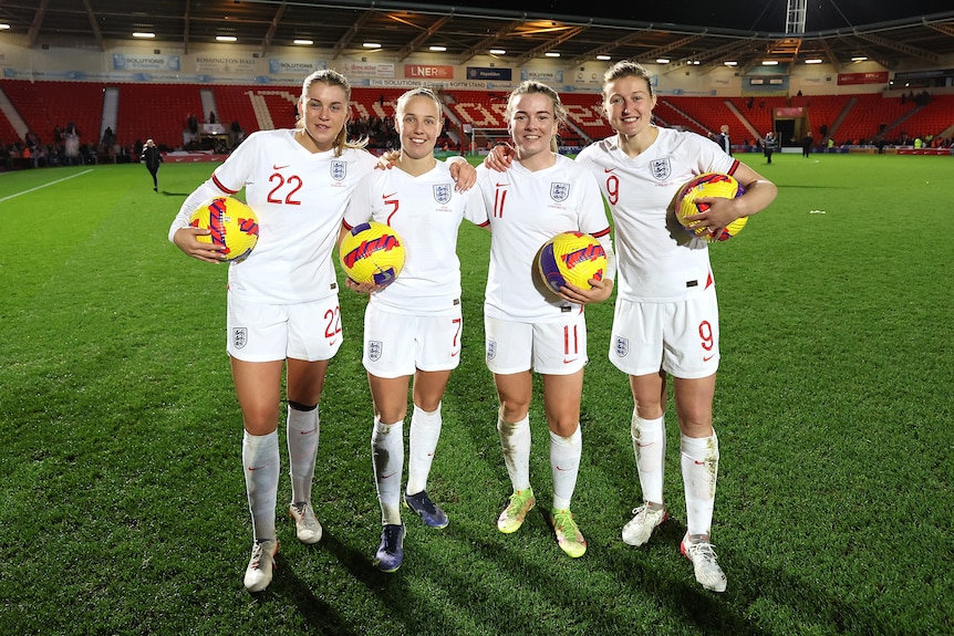 Three England women's footballers hang on to each other smiling, while holding match balls after scoring hat-tricks.