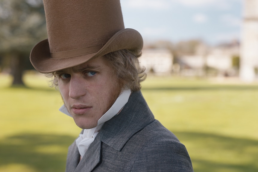 A blonde man in Regency England era tall brown top hat, white blouse shirt and grey jacket, casts a suspicious gaze to his left.