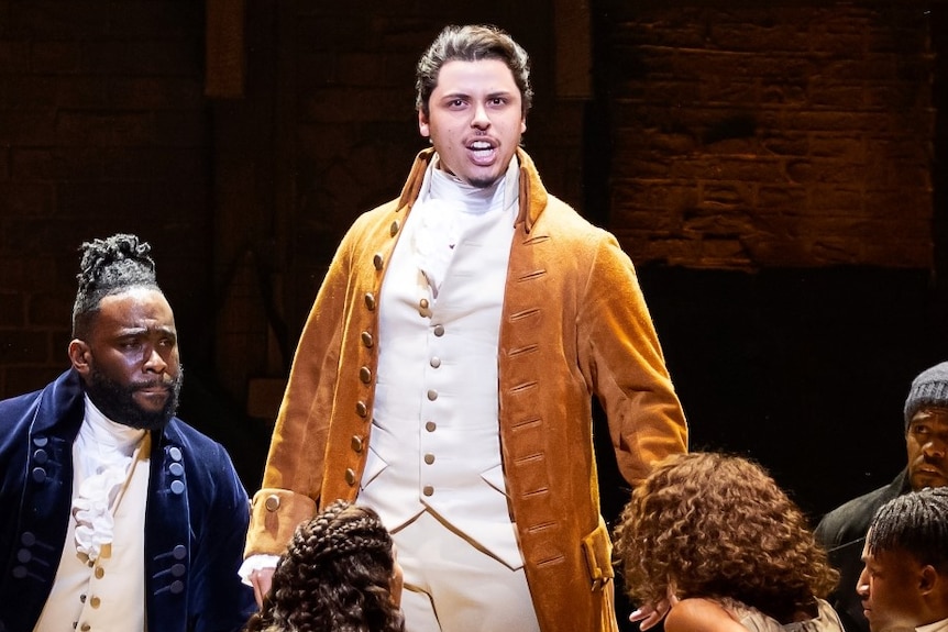 A scene from the musical Hamilton with the protagonist Alexander Hamilton standing on a box on a stage surrounded by the cast