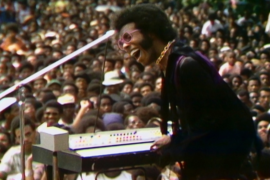 Black soul singer Sly Stone in slim-fitting black blouse and purple sunnies rocks out on keyboard in front of an animated crowd.