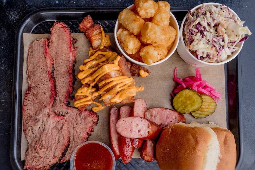 Slow cooked brisket, pork ribs, sausage, potato gems, slaw and white bread sit on a black tray.