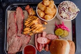 Slow cooked brisket, pork ribs, sausage, potato gems, slaw and white bread sit on a black tray.