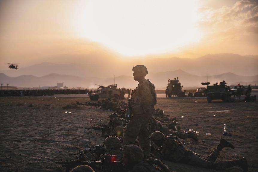 A man stands while a group of men in military gear lie on the ground with guns.