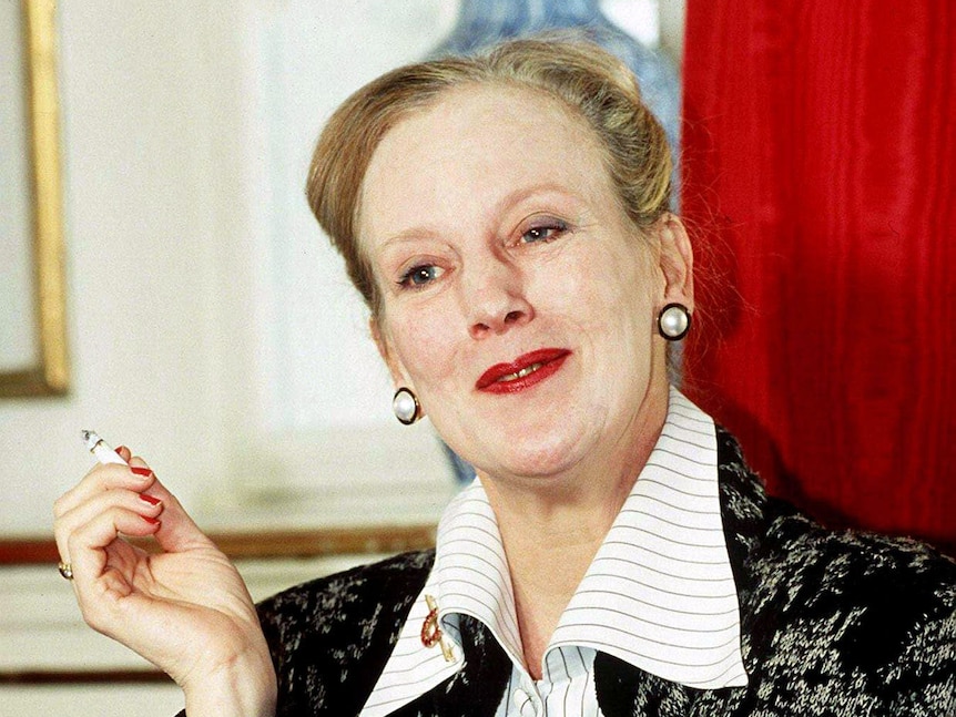 Queen Margrethe, wearing a black blazer over pin striped shirt, holds a cigarette in one hand, smiling with red lips