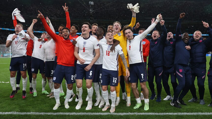 England players and coaches stand arm-in-arm and look up at the crowd while celebrating on the pitch