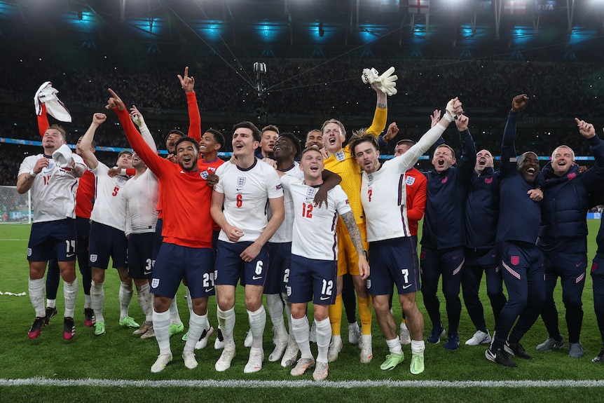 England players and coaches stand arm-in-arm and look up at the crowd while celebrating on the pitch