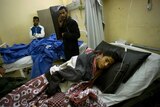 An injured boy lies in a hospital bed with men standing over him.