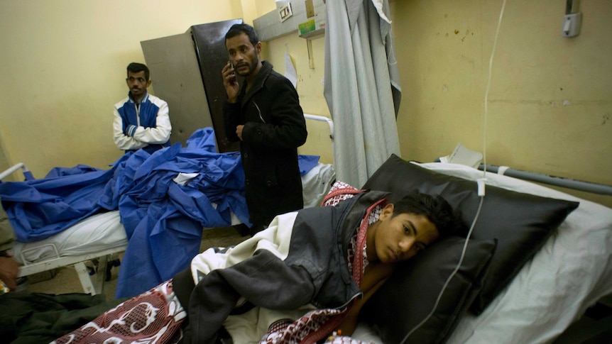 An injured boy lies in a hospital bed with men standing over him.