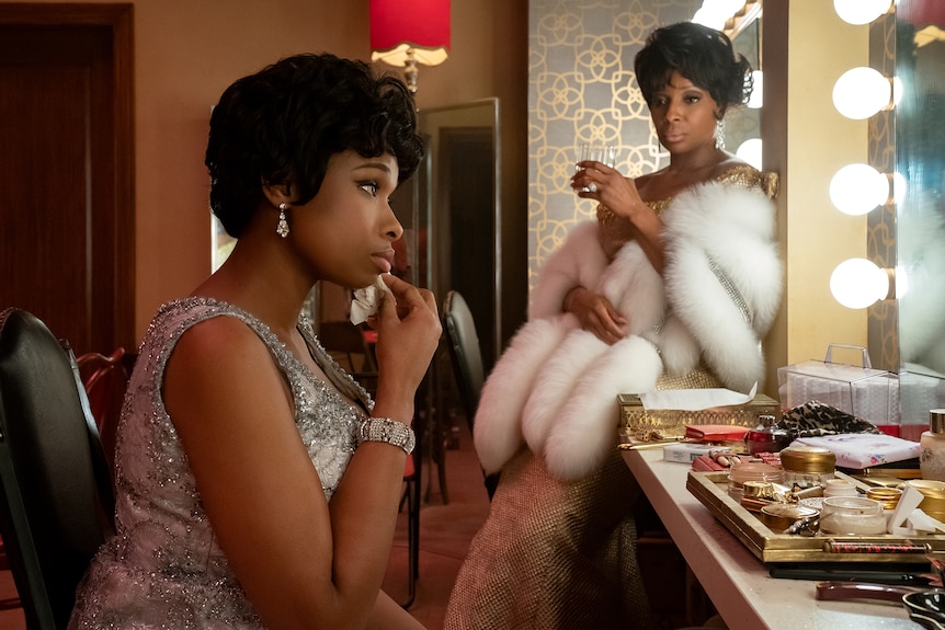 Jennifer Hudson in bedazzled gown applies makeup in a warmly lit dressing room. Mary J Blige sits beside her, wrapped in mink.