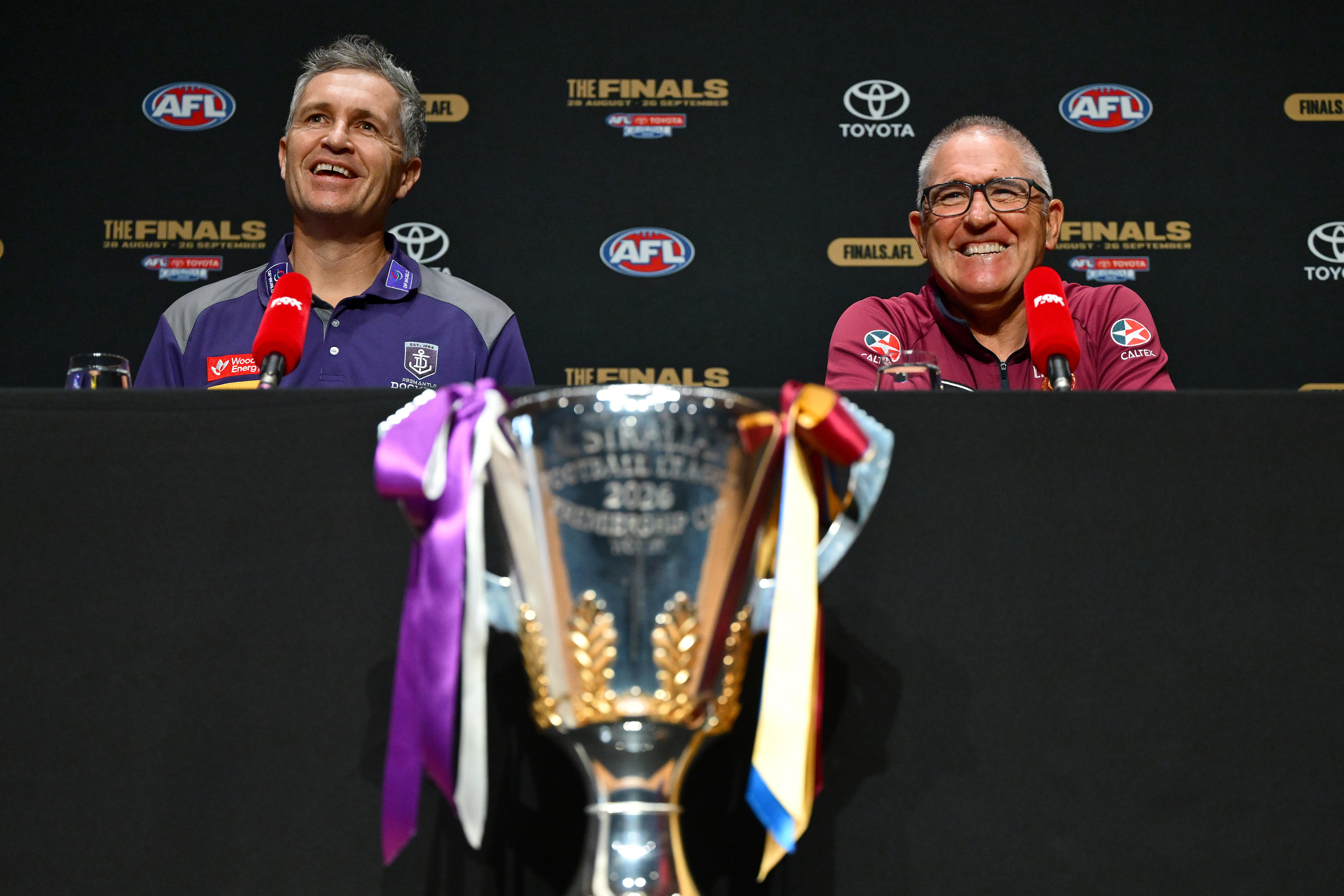Two men, one in a Fremantle polo and one in a Brisbane polo, smile during a press conference.