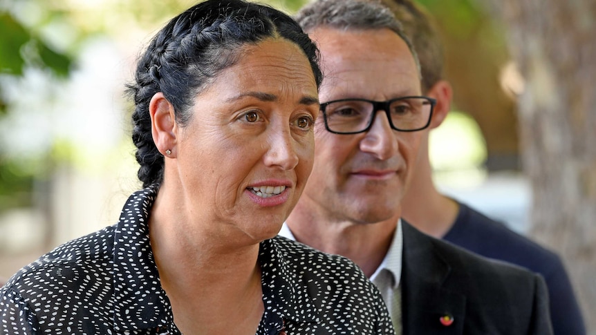 Greens Leader Richard Di Natalie is slightly out of focus as he stands behind Alex Bhathal, who is speaking at a presser.