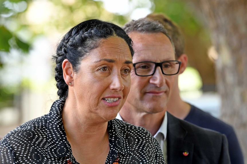 Greens Leader Richard Di Natalie is slightly out of focus as he stands behind Alex Bhathal, who is speaking at a presser.