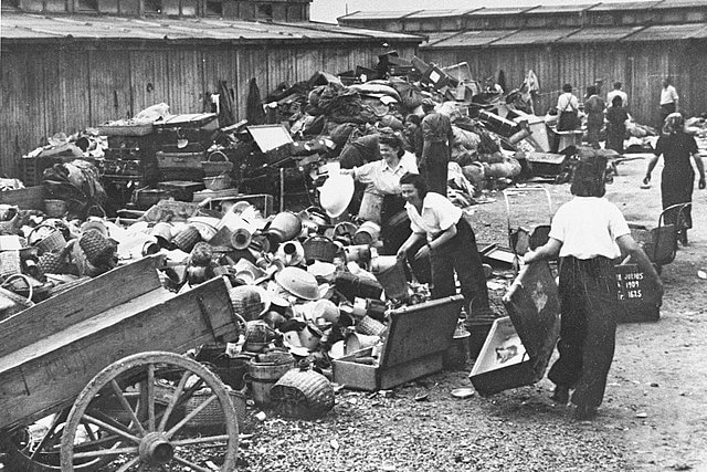 A black and white image showing women in Auschwitz sorting through towering piles of suitcases, vases and clothes.