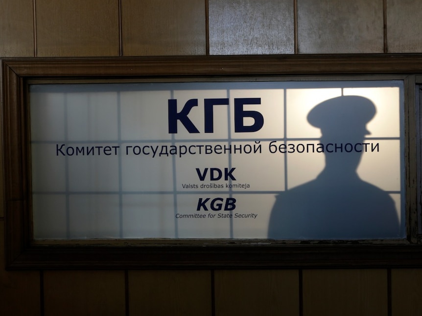 The silhouette of a man in uniform can be seen behind a window with a sign on it that says KGB on it 