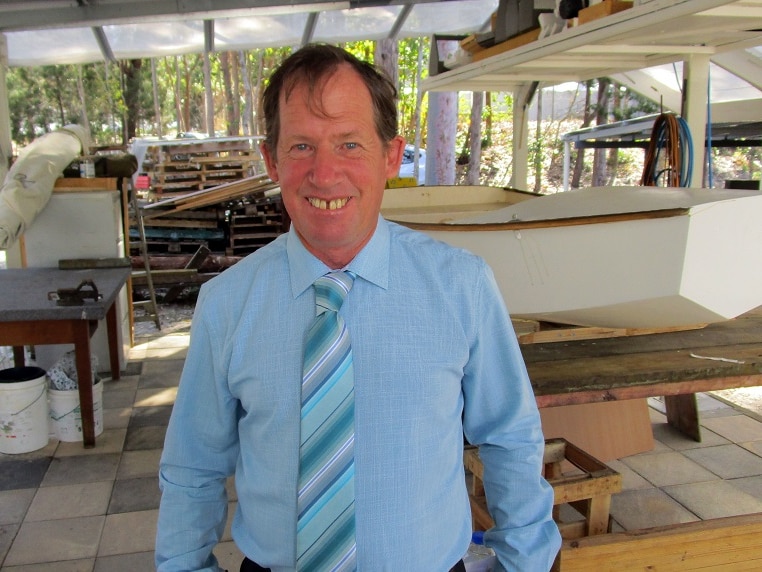 Middle aged man stands smiling at camera in an open shed with lots of wood and tinkerings in the background.