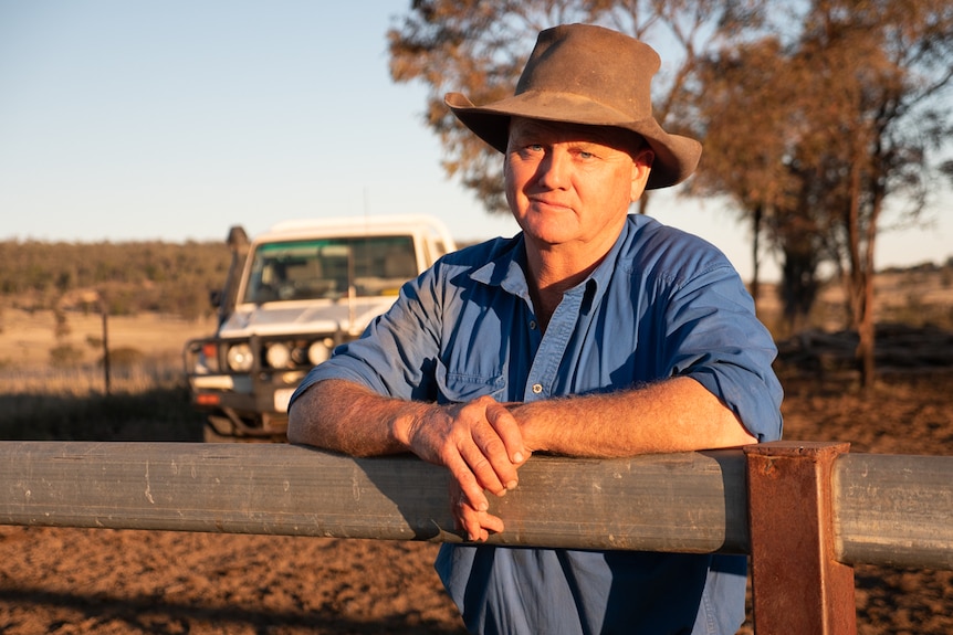 A middle-aged man in a blue shirt and brown hat leans on a farmland fence with ute and trees behind.