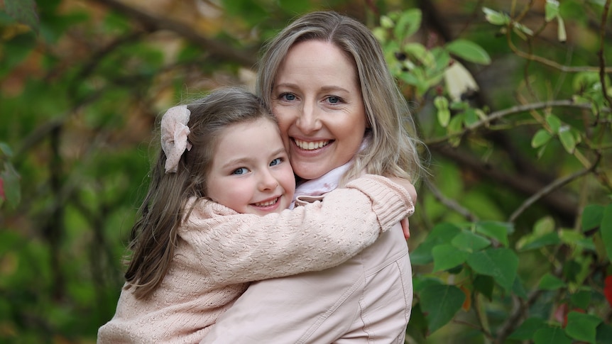 A blonde woman hugging her young daughter behind greenery.