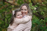 A blonde woman hugging her young daughter behind greenery.