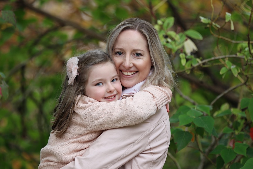 A blonde woman hugging her young daughter behind greenery.