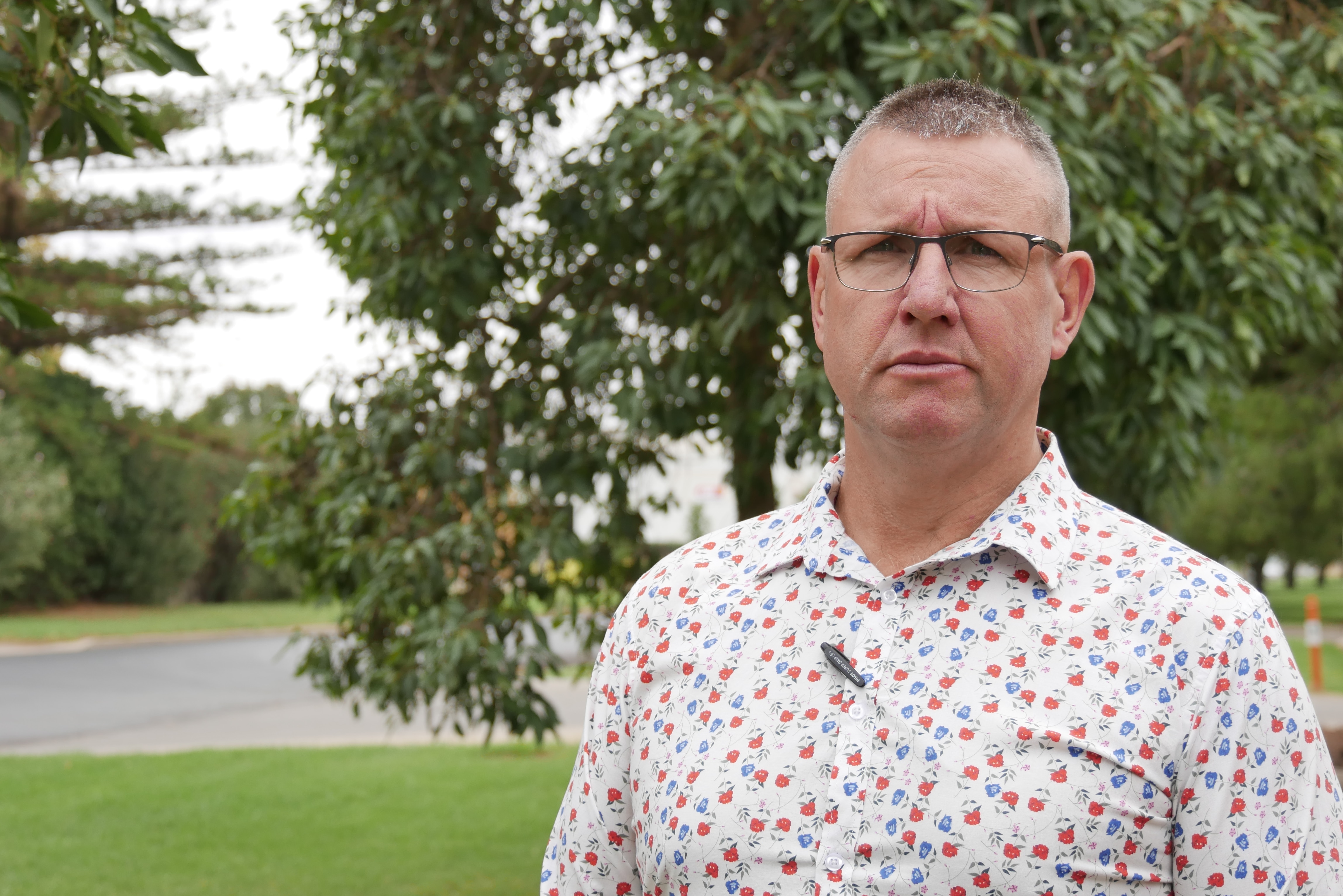 Un hombre con gafas y una camisa manchada frunce el ceño frente a un árbol