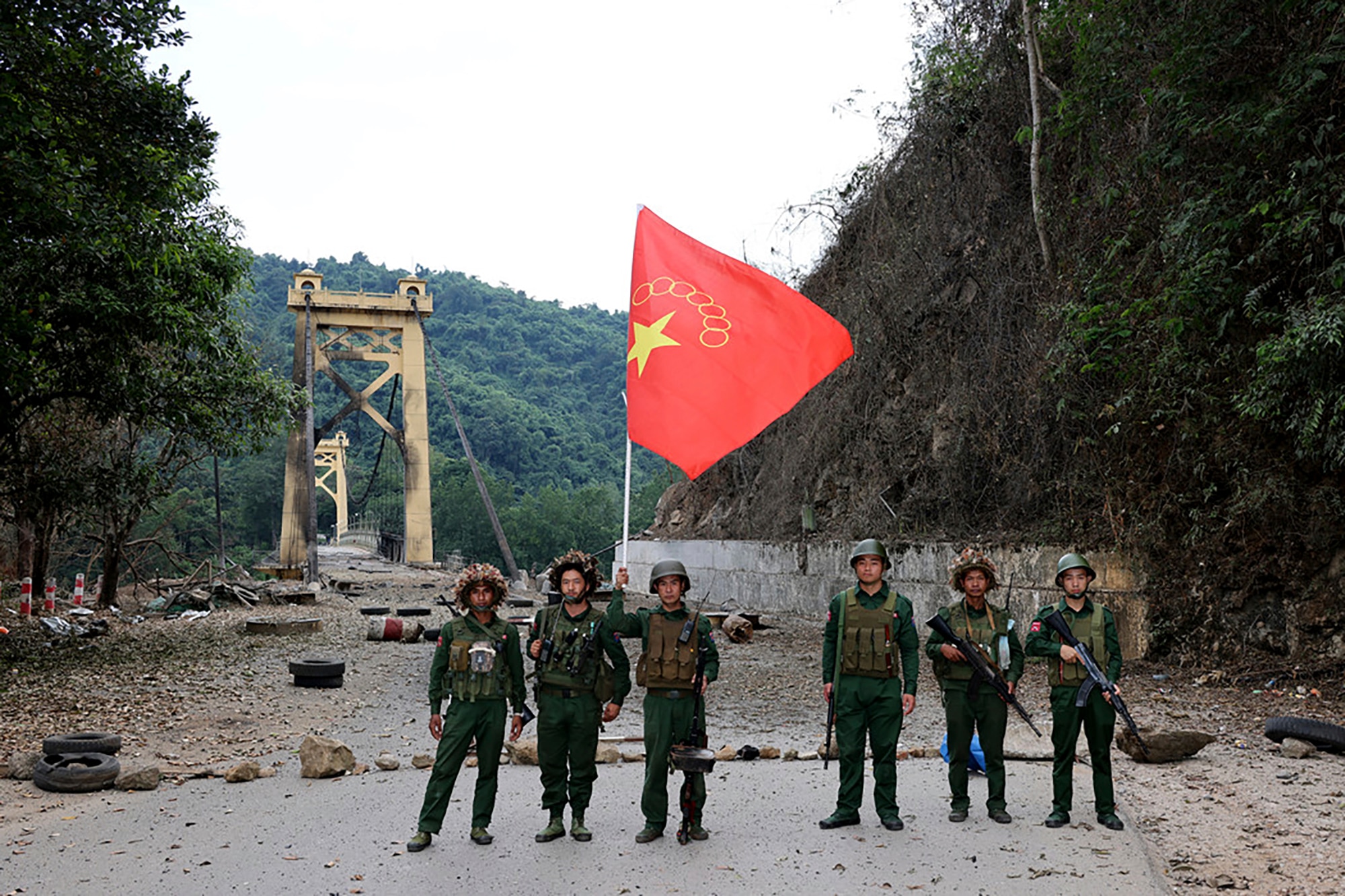 Soldiers with an MNDAA flag pose for a photo in front of a bridge.