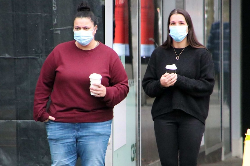 Two women wearing blue surgical masks, holding coffee on the street in Melbourne.