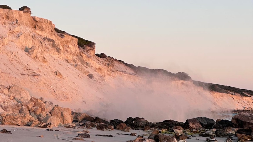 A sandy coloured cliff at sunset, with dust rising off rocks underneath.