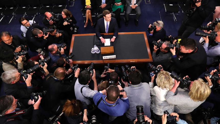 Mark Zuckerberg sits at a desk by himself, surrounded buy photographers.