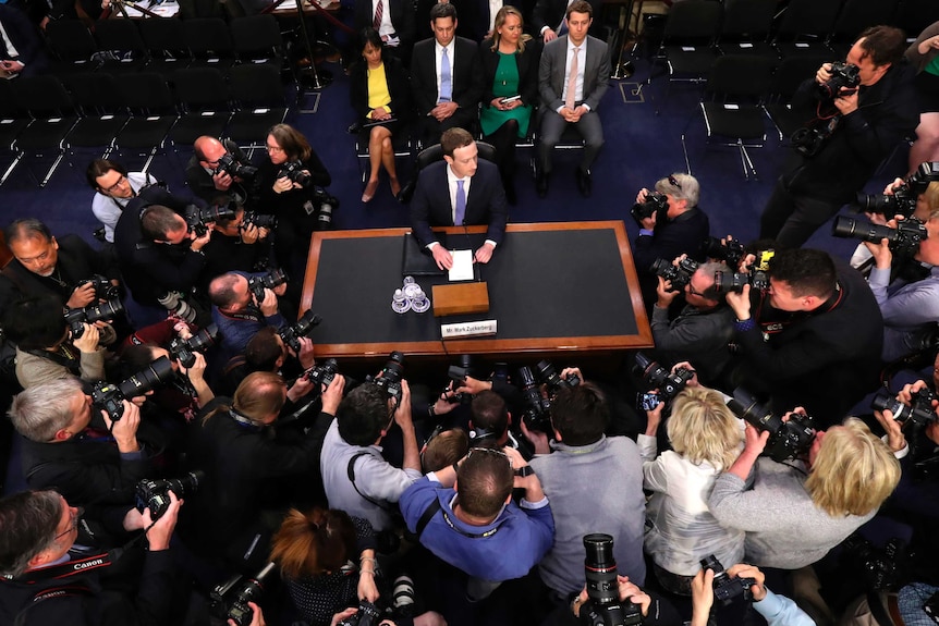Mark Zuckerberg sits at a desk by himself, surrounded buy photographers.
