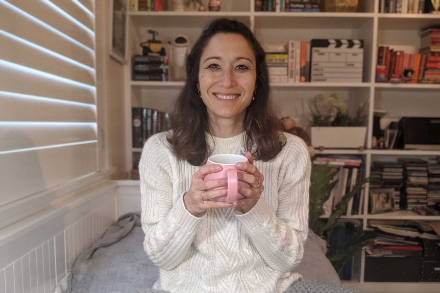 Kumi holding tea cup sitting on couch with book shelves in background.