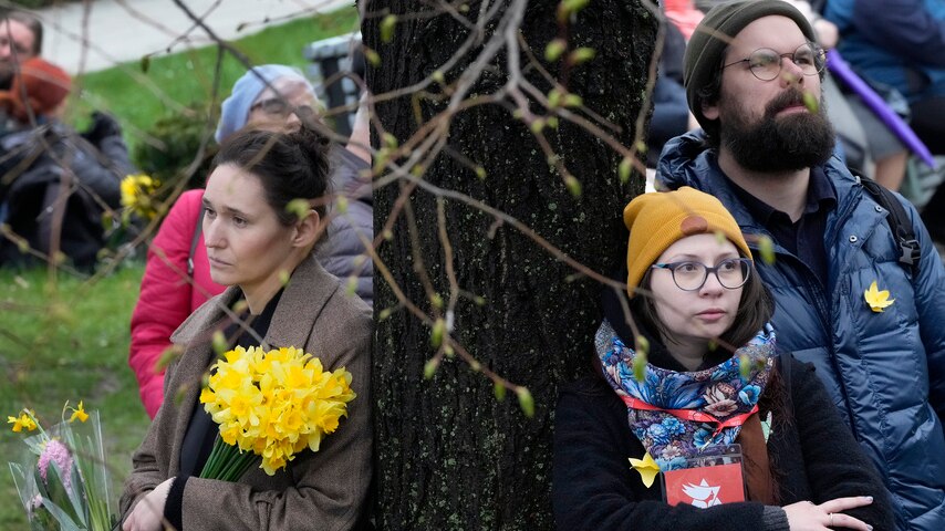 A sad-looking woman leans against a tree, holding a bunch of daffodils with another woman leaning on the tree beside her.