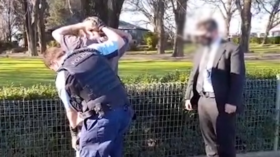 A man stands with his hands on his head on a footpath beside a park while a uniformed police officer begins to handcuff him.