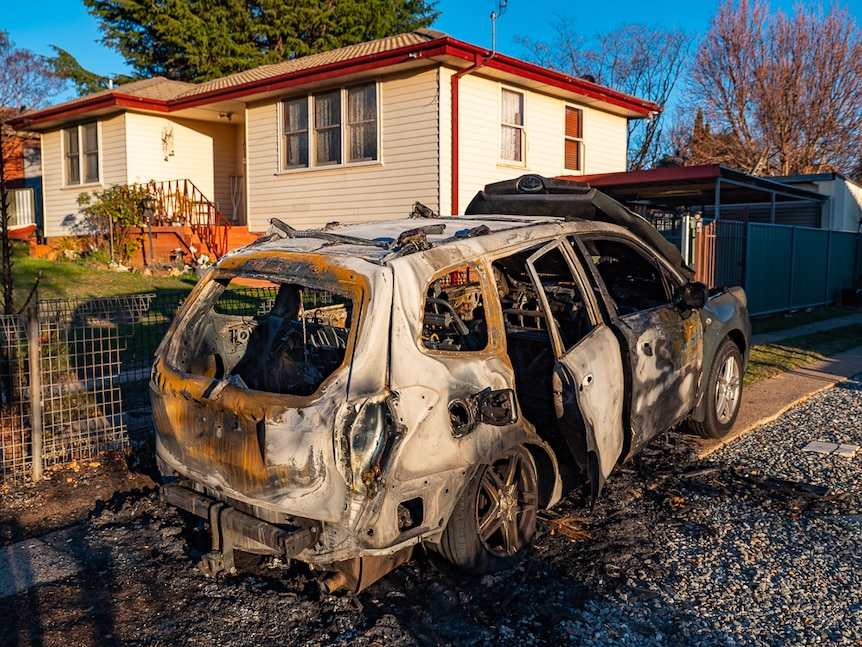 A burnt out car lies in the driveway of a suburban home.