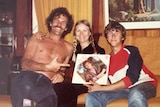 A shirtless moustached man points at an LP record on an old couch next to a woman with piggy tails and young man.