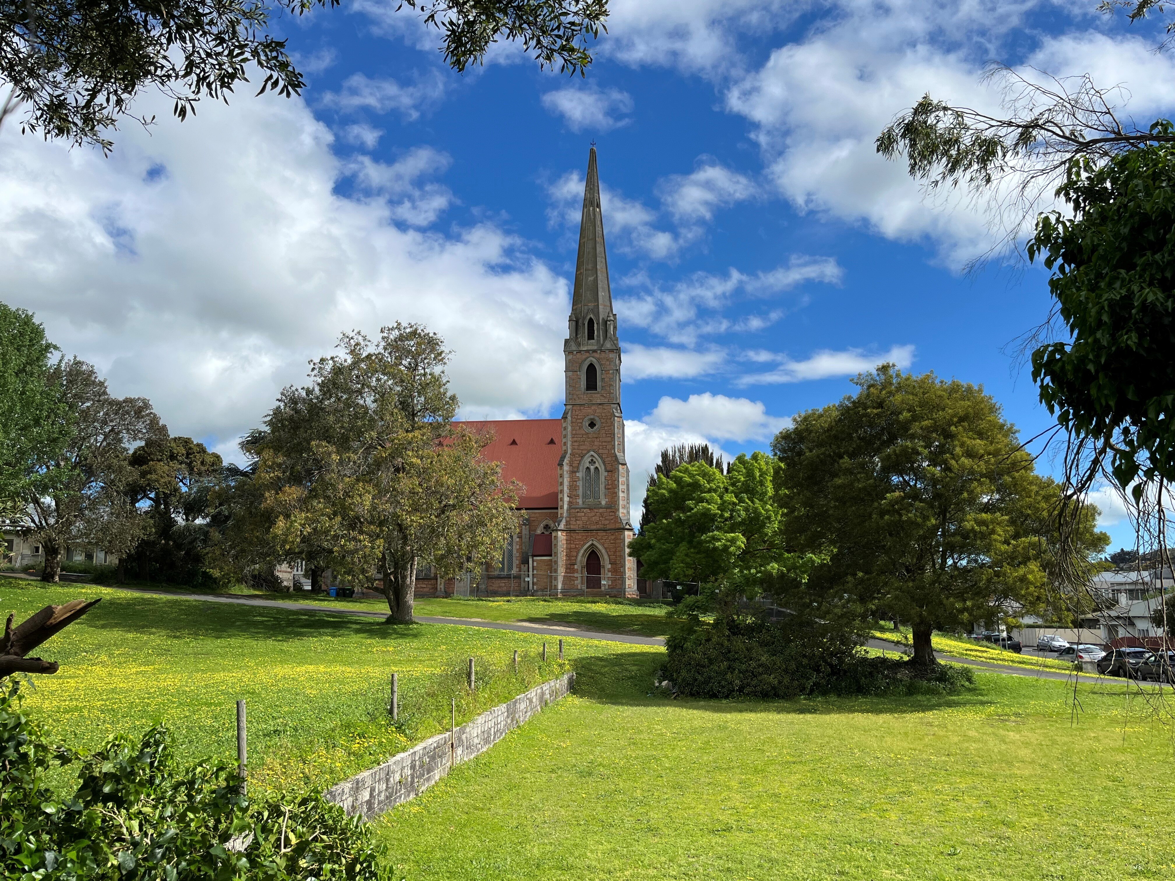 A church with a tall, narrow steeple under a bright blue sky with clouds. There are trees and green grass in the foreground.