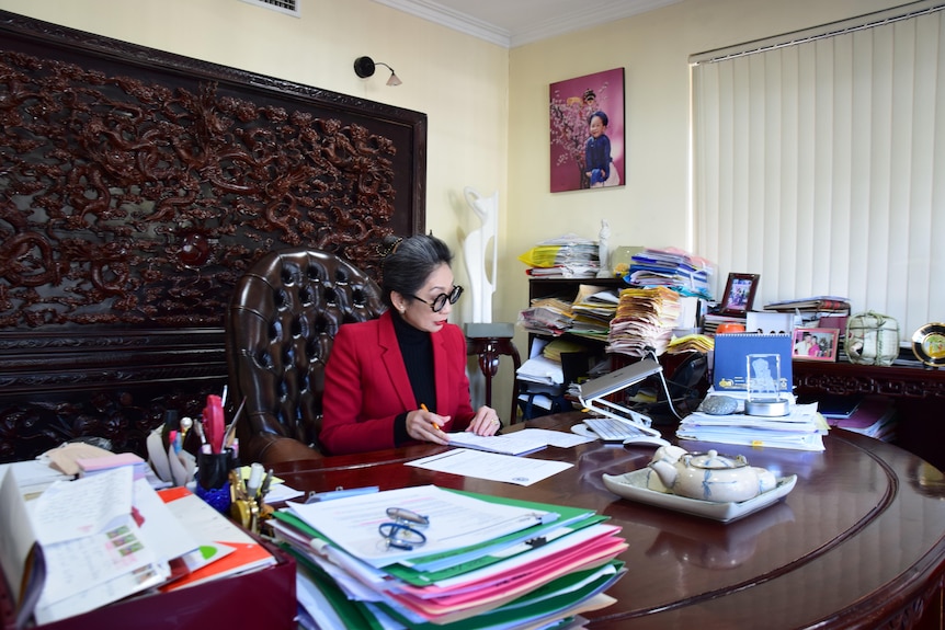 Asian woman in a black top and red blazer sitting at a large wooden desk with piles of papers beside her.