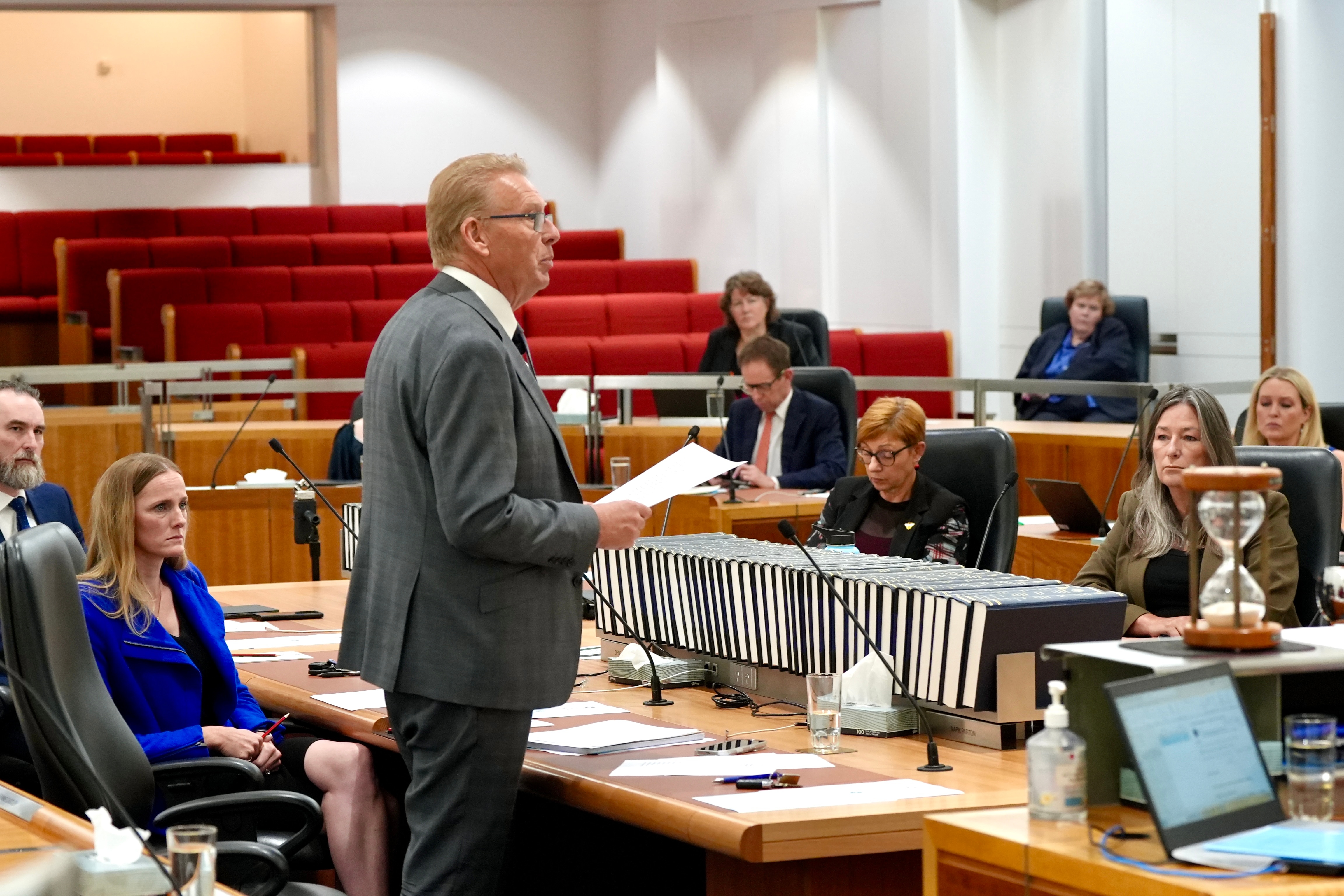 A man with blonde hair wearing a suit speaks in a room with red seats.