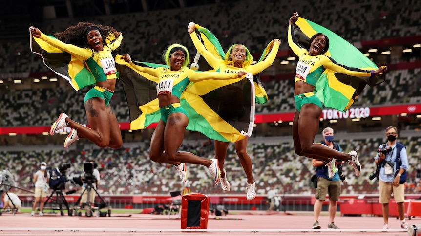 Briana Williams, Elaine Thompson-Herah, Shelly-Ann Fraser-Pryce and Shericka Jackson jump in the air holding Jamaican flags.