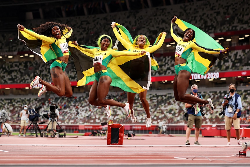 Briana Williams, Elaine Thompson-Herah, Shelly-Ann Fraser-Pryce and Shericka Jackson jump in the air holding Jamaican flags.