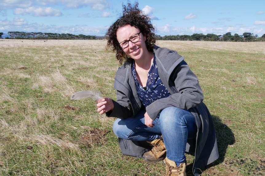 Woman kneels in paddock holding up a goose feather.