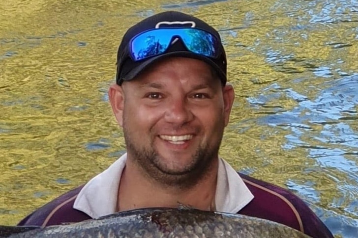 A smiling man holding a fish, wearing a cap and sunglasses pushed up on his forehead.