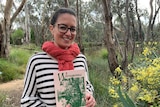 A young smiling woman, hair tied back, black-rimmed glasses, melon-coloured scarf holds a book in a bush park.