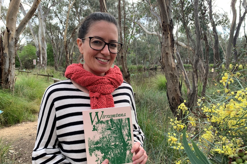 A young smiling woman, hair tied back, black-rimmed glasses, melon-coloured scarf holds a book in a bush park.