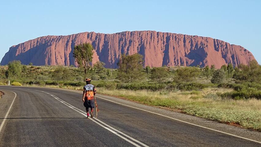 Clinton walks on a road with Uluru in the background.